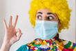 © AliciaFdez - Linda mujer joven sonriendo mientras está vestida con un disfraz de payaso con una mascarilla quirúrgica desechable y una peluca de payaso amarilla. Protección contra el virus sars-cov-2