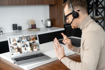 Young fit businessman in headset using app for distance video communication with coworkers, meeting online in pandemic, looking at laptop screen with people profiles, saying hello, showing two hands