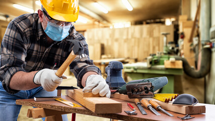 Wall Mural - Carpenter worker at work in the carpentry workshop, wears helmet, goggles, leather gloves and surgical mask to prevent coronavirus infection. Preventing Pandemic Covid-19 at the workplace.