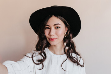 Curly chinese woman taking selfie. Front view of asian model in hat posing on beige background.