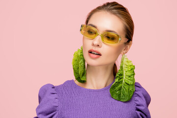 stylish woman wearing colored eyeglasses and earrings made of fresh lettuce isolated on pink