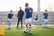 © matimix - Young Boy Training Sports in Sunny Summer Day. Happy Kids Practicing Soccer on Schoolyard. Young Coach With Children Football Players on Training Session. Sports Stadium Seats in the Background