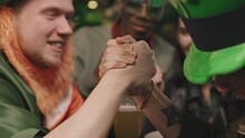 Panning Closeup With Slowmo Of Joyful Man In Green Irish Hats Arm Wrestling While Sitting At Table In Local Pub And Their Friends Watching, Everyone Having Fun