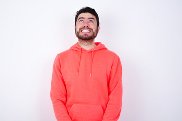 Wall Mural - Portrait of mysterious young Caucasian bearded man wearing pink hoodie against white wall looking up with enigmatic smile. Advertisement concept.