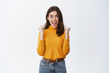 Yes victory. Excited girl screams and celebrated success, raising hands in fist pump and smiling amazed at camera, achieve goal or win prize, standing happy on white background