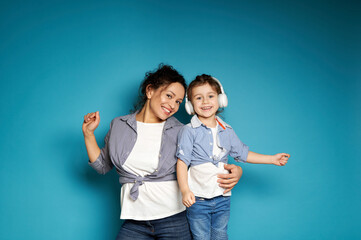 Adorable baby girl with headphones dancing near her mother hugging her on a blue background with copy space