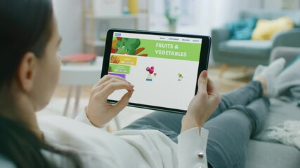 Wall Mural - Woman Lying on Couch Uses Touchscreen Tablet Computer Browsing through Online Food Delivery Market, Choosing Through Vegetables Section, Fresh Goods. Internet Store Software with Food to Order