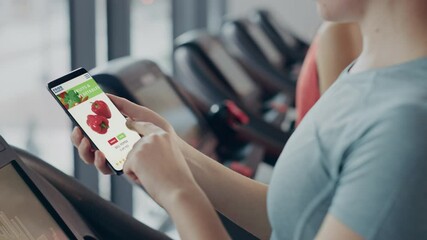 Wall Mural - In Gym: Beautiful Woman Running on Treadmill Uses Smartphone App to Order Healthy Food from Online Delivery Store, Browsing Through Vegetables, Fresh Goods Section. Internet Product Market Software