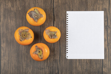 Four ripe persimmon fruits and empty notebook on wooden surface