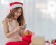 © Bangkok Click Studio - A young long brown hair female wearing red and white christmas hat and pink sleeveless dress using her silver mobile phone surrounded by gift boxes in front of white curtain background
