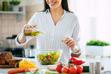 Wall Mural - Close up photo of a smiling young woman makes a fresh vegan salad while she uses olive oil for it.