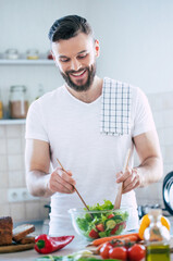 Handsome happy bearded man is preparing wonderful fresh vegan salad in the kitchen at home