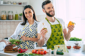 Wall Mural - Excited smiling young couple in love making a super healthy vegan salad with many vegetables in the kitchen and having fun