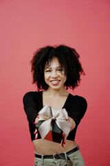 Wall Mural - Shows holding a black gift box with a bow. young woman of Afro appearance, curly lush hair. She is dressed in a black comfortable jacket. He looks at the camera and smiles. Copy space.