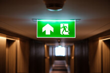 Emergency Exit Sign At The Corridor In Building. Green Fire Exit Sign Hanging On Ceiling On Dark Corridor In Building Near Fire Emergency Exit Door. Green Emergency Exit Sign.