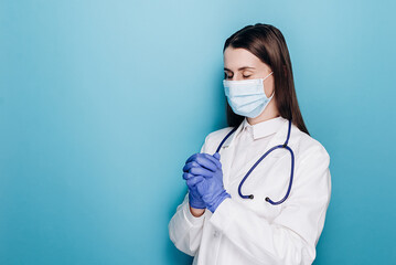 Young woman doctor in white medical gown sterile face mask gloves holding hands folded in prayer, isolated on blue background. Epidemic pandemic coronavirus 2019-ncov sars covid-19 flu virus concept