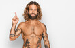 Handsome man with beard and long hair standing shirtless showing tattoos showing and pointing up with finger number one while smiling confident and happy.