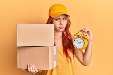 Poster - Young redhead woman holding delivery package and alarm clock skeptic and nervous, frowning upset because of problem. negative person.