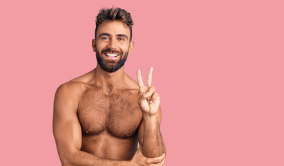 Wall Mural - Young hispanic man wearing swimwear shirtless smiling with happy face winking at the camera doing victory sign. number two.