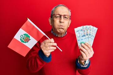 Sticker - Handsome senior man with grey hair holding peru flag and peruvian sol banknotes puffing cheeks with funny face. mouth inflated with air, catching air.