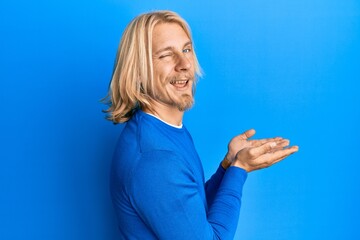 Canvas Print - Caucasian young man with long hair presenting with open palms, holding something winking looking at the camera with sexy expression, cheerful and happy face.