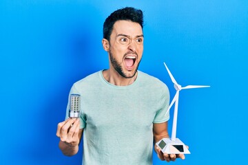 Poster - Young hispanic man holding windmill and led lightbulb angry and mad screaming frustrated and furious, shouting with anger. rage and aggressive concept.