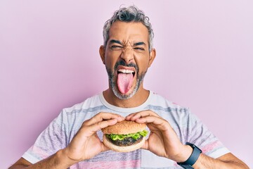 Sticker - Middle age grey-haired man eating a tasty classic burger sticking tongue out happy with funny expression.