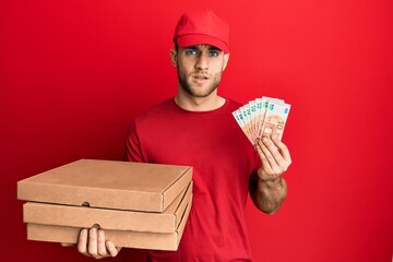 Canvas Print - Young caucasian man holding take away food and 10 euros banknotes in shock face, looking skeptical and sarcastic, surprised with open mouth