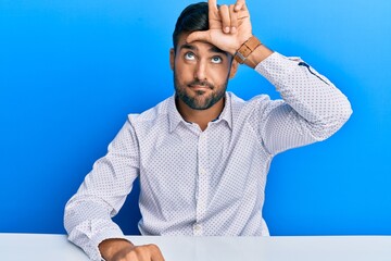 Sticker - Handsome hispanic man wearing business clothes sitting on the table making fun of people with fingers on forehead doing loser gesture mocking and insulting.