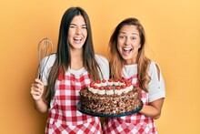 Hispanic Family Of Mother And Daughter Wearing Baker Apron Holding Homemade Cake Smiling And Laughing Hard Out Loud Because Funny Crazy Joke.