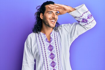 Poster - Young handsome man with long hair wearing bohemian and hippie shirt very happy and smiling looking far away with hand over head. searching concept.