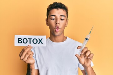 Canvas Print - Young handsome african american man holding botox banner and needle making fish face with mouth and squinting eyes, crazy and comical.