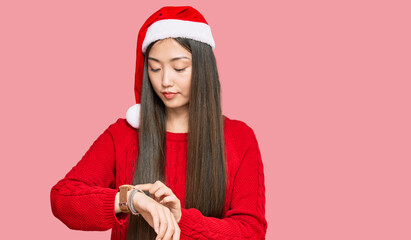 Poster - Young chinese woman wearing christmas hat checking the time on wrist watch, relaxed and confident