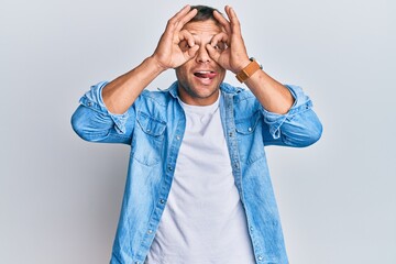 Wall Mural - Handsome muscle man wearing casual denim jacket doing ok gesture like binoculars sticking tongue out, eyes looking through fingers. crazy expression.