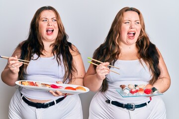 Wall Mural - Plus size caucasian sisters woman eating sushi using chopsticks angry and mad screaming frustrated and furious, shouting with anger. rage and aggressive concept.