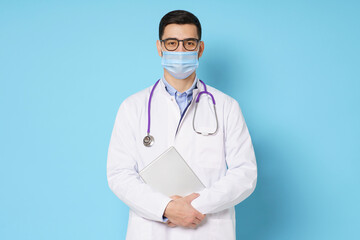 Wall Mural - Young male doctor wearing medical mask, glasses and stethoscope round his neck, holding tablet, isolated on blue background