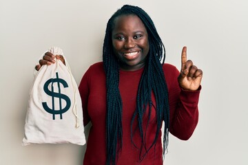 Poster - Young black woman with braids holding dollars bag smiling with an idea or question pointing finger with happy face, number one