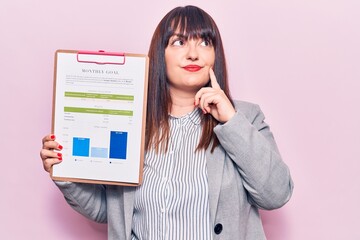 Wall Mural - Young plus size woman holding clipboard with chart information paper serious face thinking about question with hand on chin, thoughtful about confusing idea