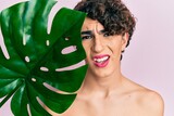 Fototapeta  - Young man wearing woman make up holding green plant leaf close to beautiful face clueless and confused expression. doubt concept.