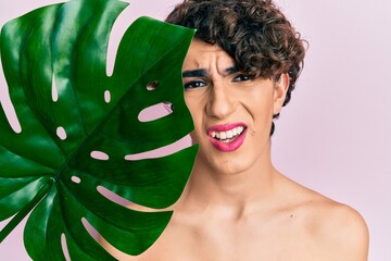 Young man wearing woman make up holding green plant leaf close to beautiful face clueless and confused expression. doubt concept.