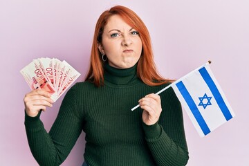 Sticker - Beautiful redhead woman holding 20 shekels banknotes and israel flag skeptic and nervous, frowning upset because of problem. negative person.