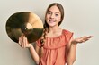 Beautiful brunette little girl holding golden cymbal plates celebrating achievement with happy smile and winner expression with raised hand