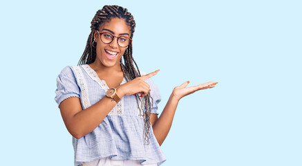 Sticker - Young african american woman with braids wearing casual summer clothes and glasses amazed and smiling to the camera while presenting with hand and pointing with finger.