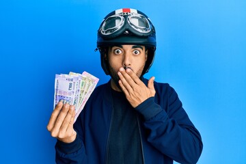 Canvas Print - Young hispanic man wearing motorcycle helmet holding indian rupee covering mouth with hand, shocked and afraid for mistake. surprised expression