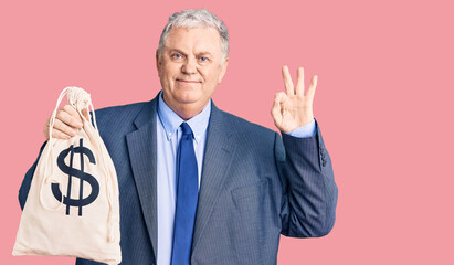 Sticker - Senior grey-haired man wearing business suit holding money bag with dollar symbol doing ok sign with fingers, smiling friendly gesturing excellent symbol