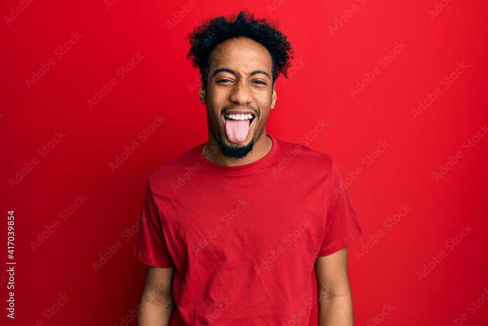 Young african american man with beard wearing casual red t shirt sticking tongue out happy with funny expression. emotion concept.