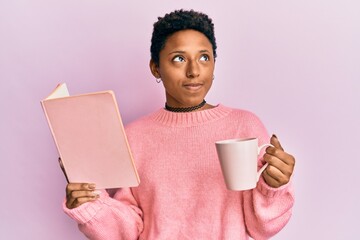 Wall Mural - Young african american girl reading a book and drinking a cup of coffee smiling looking to the side and staring away thinking.