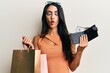 Beautiful hispanic woman holding shopping bags and wallet with dollars making fish face with mouth and squinting eyes, crazy and comical.