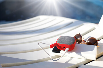 Red music portable speaker is charged from the power bank via usb on a deck chair near the pool. Concept is always in touch, travel gadgets, external battery for a smartphone. Place for text. Flatlay