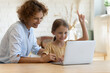 © fizkes - I know. Supportive mother watch little daughter do homework online on laptop raise finger up offer correct answer. School age girl sit by pc show eureka gesture share good idea with elder grown sister
